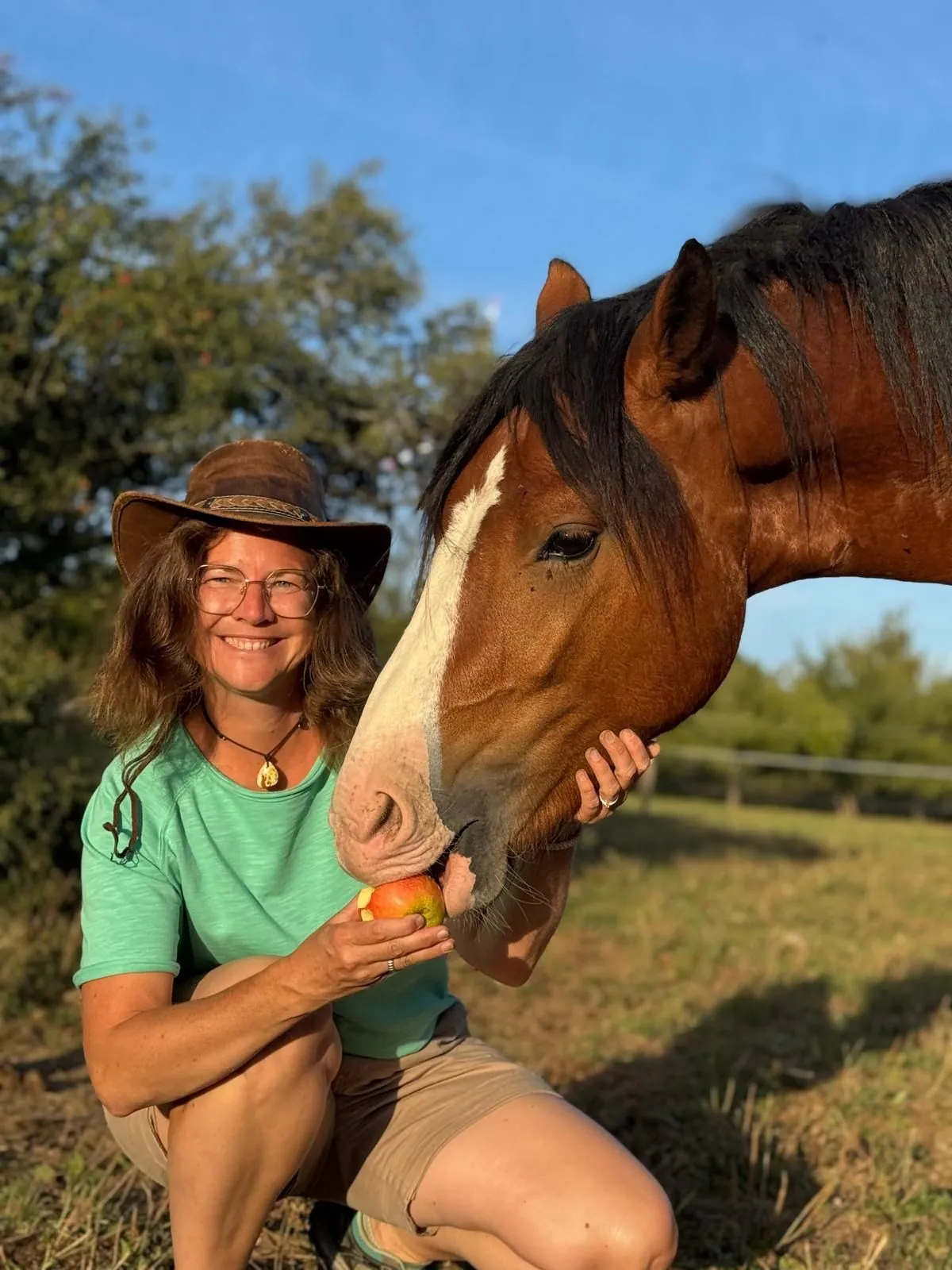 Christina mit Hut und Brille hockt lächelnd auf der Weide und füttert ihr braunes Pferd mit Blesse mit einem Apfel