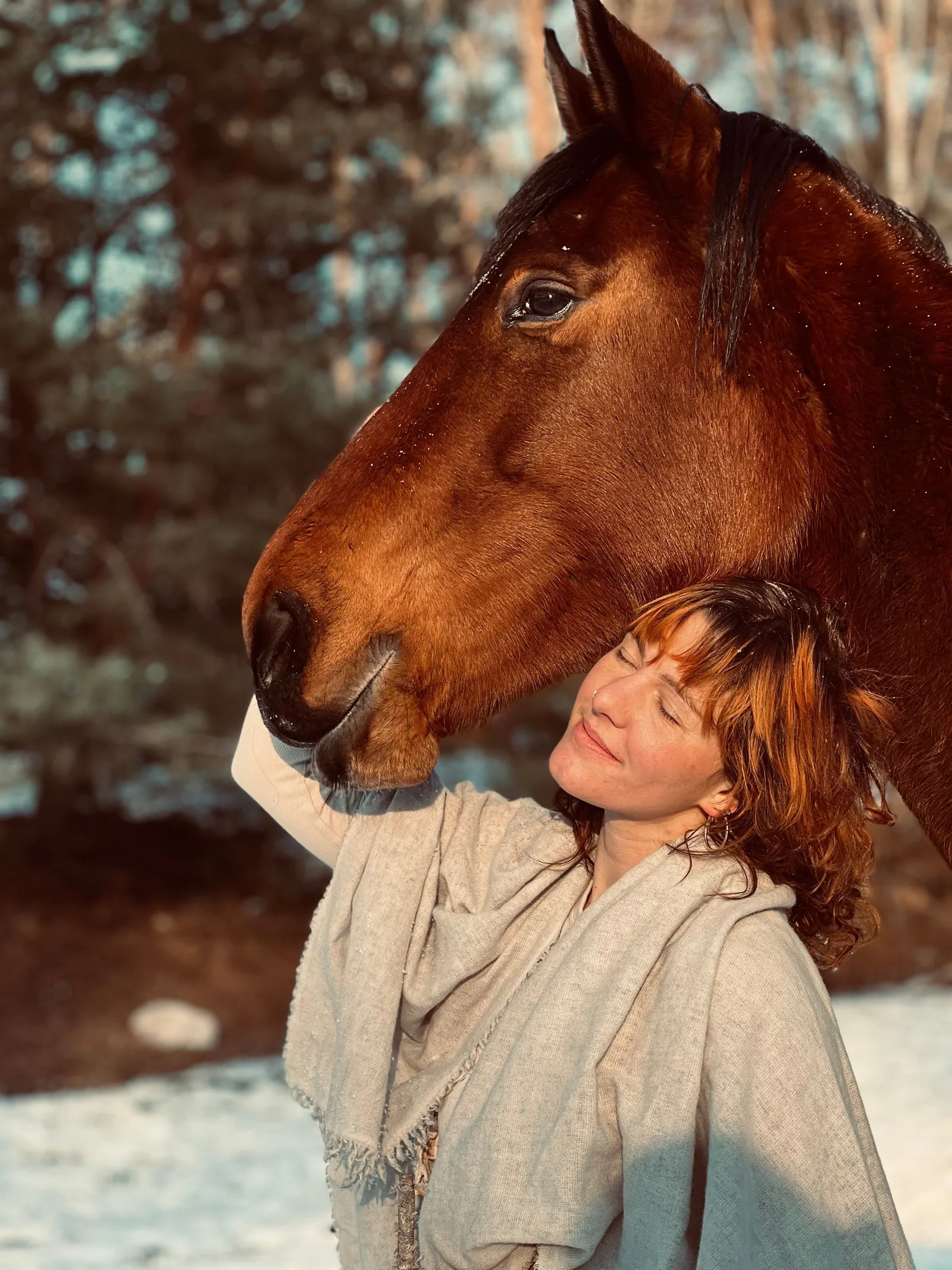 Zoe Fröhlich lehnt mit geschlossenen Augen den Kopf an ihr braunes Pferd, im Hintergrund Schnee und Bäume in der Wintersonne