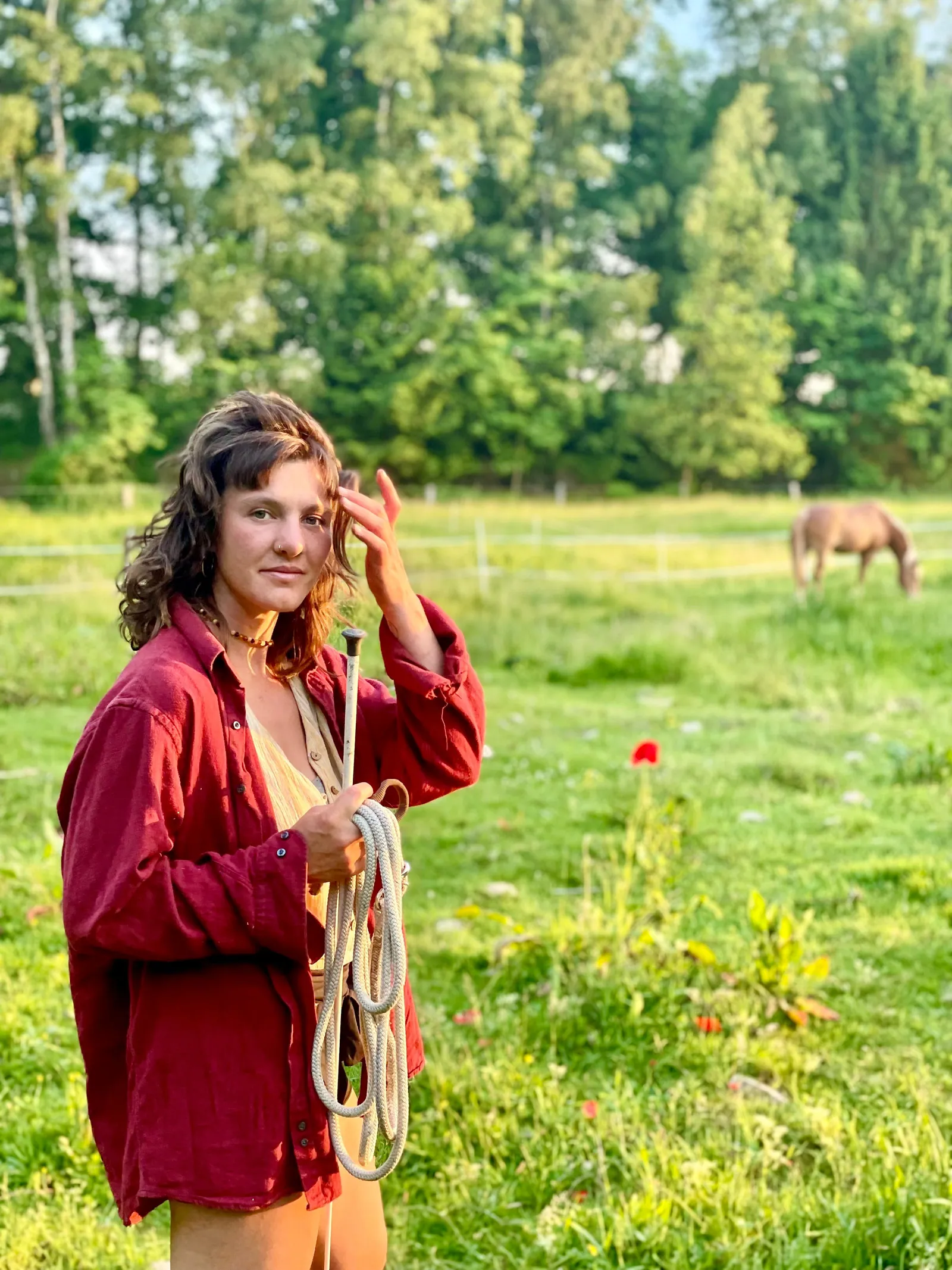 Zoe Fröhlich im roten Hemd mit aufgerolltem Seil und Stick auf einer Sommerwiese, im Hintergrund grast ein Pferd