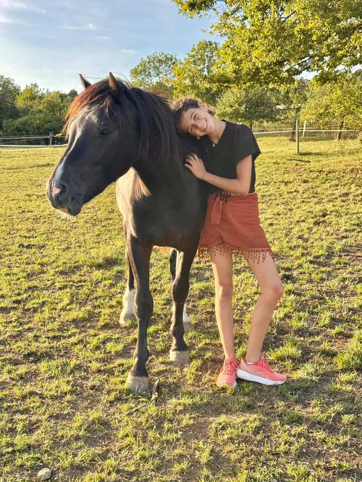 Sammy lehnt lächelnd den Kopf an den Hals ihres dunklen Pferdes, die beiden stehen auf einer sonnigen Weide